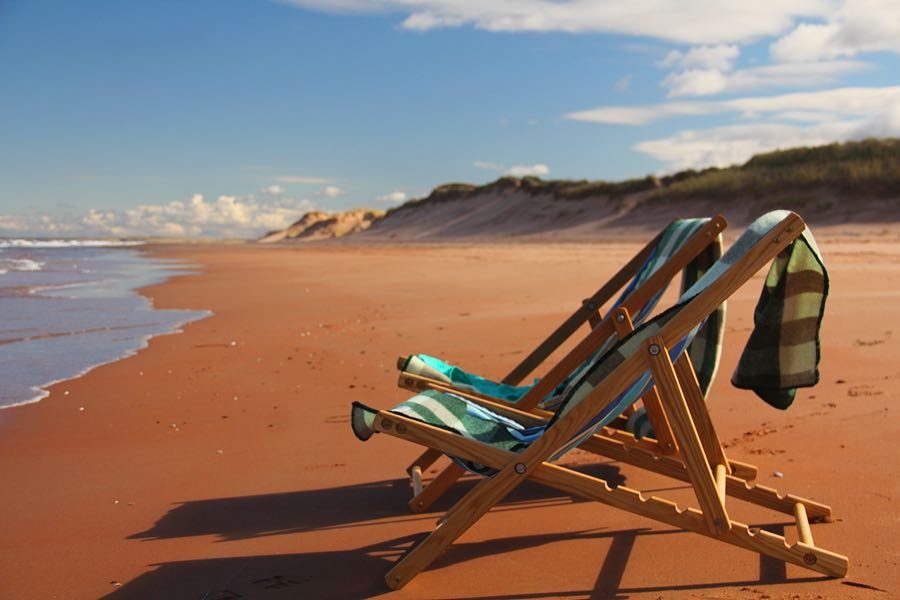 Two wooden deck chairs with towels over them on the red sand, looking down a long empty beach
