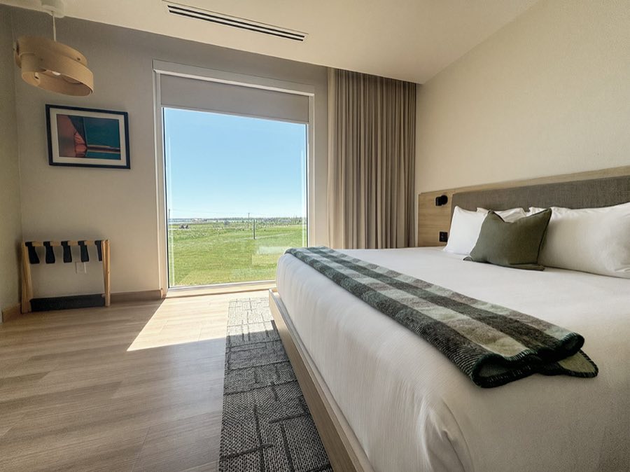 A guest room at Blackbush, bed made up with a green plaid throw and a floor to ceiling window looking out over open field to the water